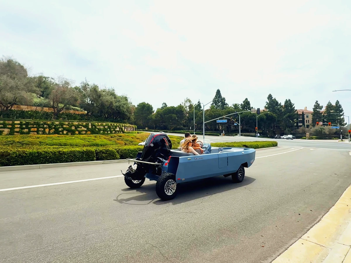 A blue WaterCar being driven on a dry paved road, with a seated helm driver, lowered road wheels and the complete hull and outboard visible.